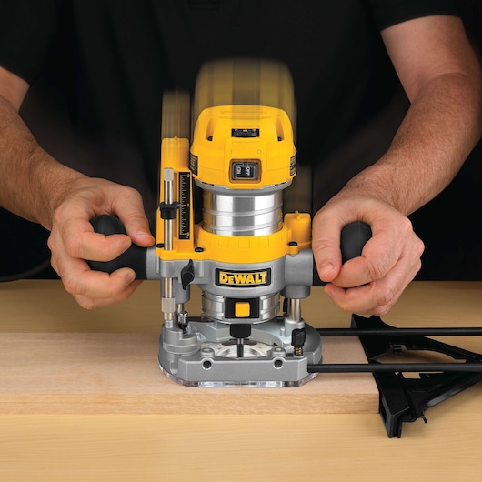 A close-up of a person using a DEWALT D26203 plunge router on a piece of wood, with both hands gripping the handles and the tool in operation.