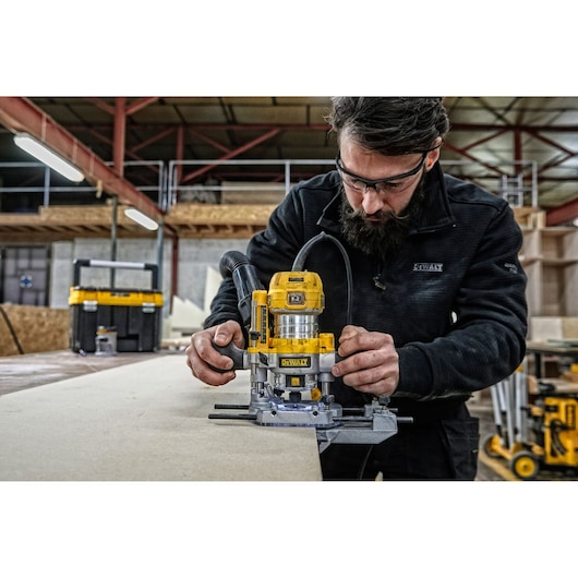 A person using a yellow DEWALT D26204K router tool on a piece of wood in a workshop. The individual's face is obscured, and there are various woodworking equipment and storage boxes in the background.