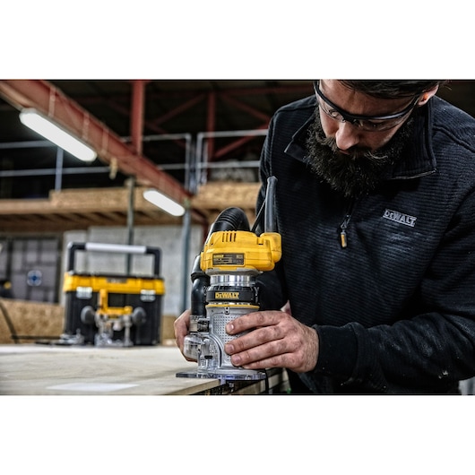 A person using a DEWALT D26204K router tool on a wooden surface in a workshop, with the person's face blurred for privacy. There is another DEWALT tool visible in the background.