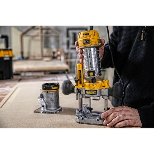 A person is using a DEWALT D26204K router tool on a piece of wood in a workshop. Another base attachment is visible on the table. The focus is on the yellow and silver DEWALT router being operated.