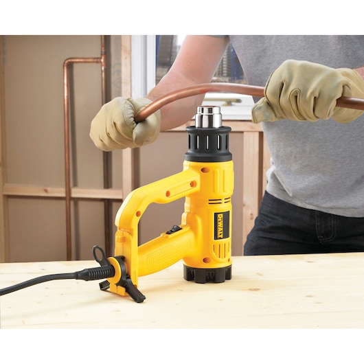 A person wearing gloves uses a DEWALT heat gun to bend a copper pipe on a wooden work surface.