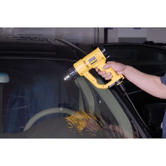 A person using a DEWALT heat gun (model D26411) to apply heat on the windshield of a car.