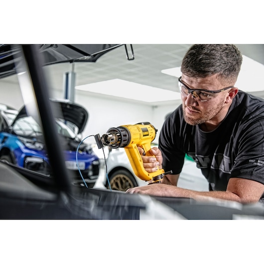 A person uses a DEWALT D26414 yellow heat gun in a garage or workshop, with cars visible in the background. The person’s face is blurred for privacy.