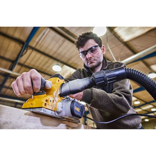 A person using a DEWALT electric planer (model D26500K) on a piece of wood in a workshop, with a dust extraction hose attached. The person's face is blurred.