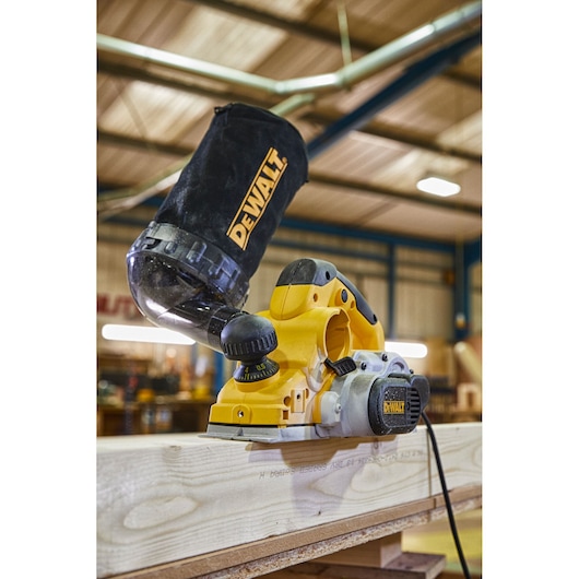 A close-up of a DEWALT electric hand planer being used on a wooden plank in a workshop. The planer has a dust collection bag attached, and the workspace background is blurred.