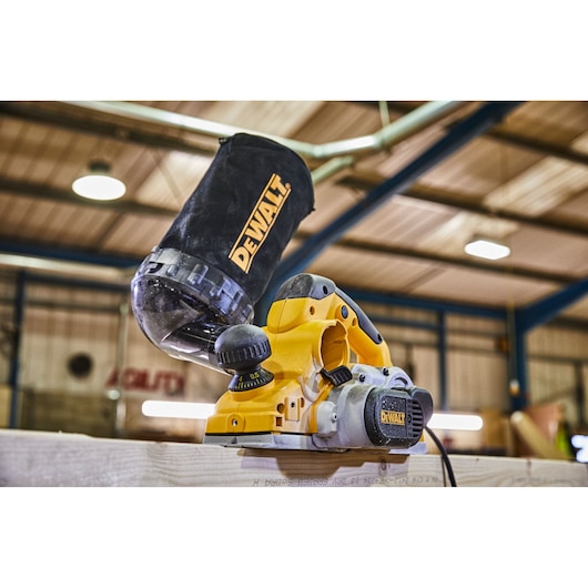 A DEWALT electric planer with a dust collection bag attached, shown in use on a wooden plank in a workshop setting.