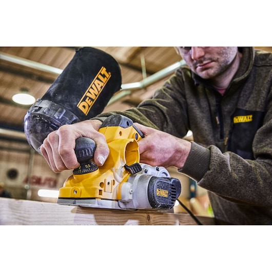 A person using a DEWALT electric planer, model D26500K, to smooth a piece of wood. The planer is yellow and black, with DEWALT branding visible, and the scene is set in a workshop environment. The person's face is blurred for privacy.