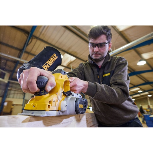 A person using a DEWALT D26500K electric planer on a wooden board in an indoor workshop. The person's face is blurred for privacy. The DEWALT branding is visible on the tool and on the person's clothing.