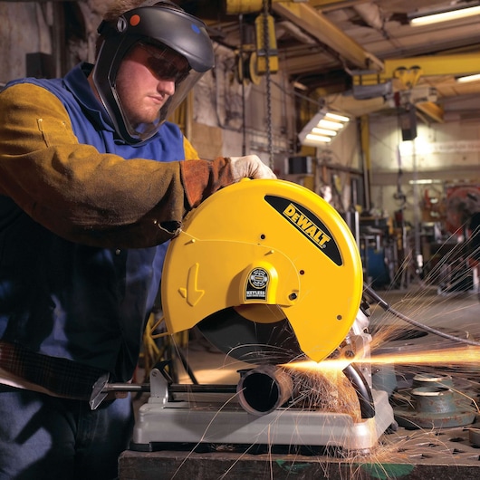 A worker wearing protective gear operates a DEWALT D28715 metal chop saw, cutting a metal pipe and producing sparks in an industrial workshop setting.