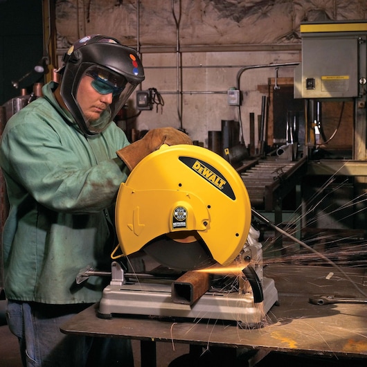 A person wearing protective gear is using a DEWALT chop saw (SKU D28715) to cut a metal pipe in an industrial workshop. Sparks are visible as the saw operates.