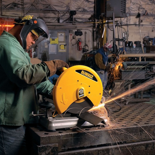 A worker wearing protective gear operates a yellow DEWALT D28715 metal cutting saw, producing sparks as it cuts through a piece of metal in an industrial workshop.
