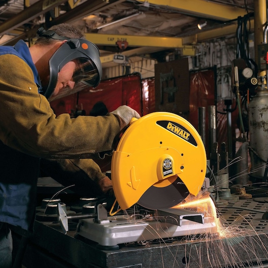 A person wearing protective gear operates a DEWALT D28715 abrasive chop saw, cutting metal and creating sparks, in an industrial workshop setting.