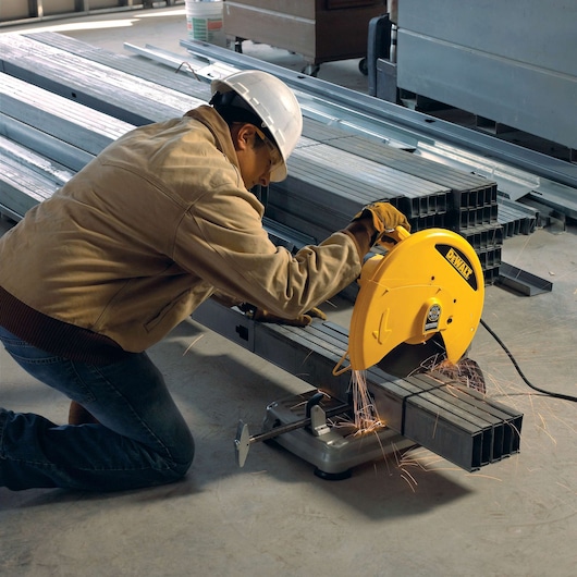A person wearing safety gear uses a DEWALT metal cutting saw to cut steel pipes in an industrial setting. Sparks are visible as the saw is in operation.