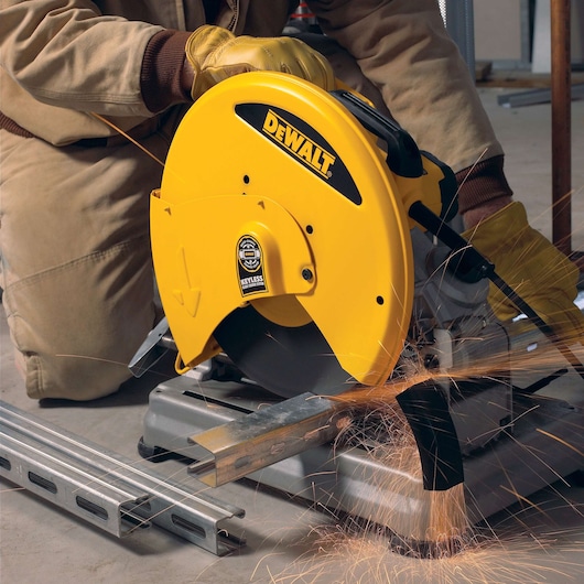 A person wearing protective gloves operates a yellow DEWALT metal cutting saw, producing sparks while cutting a steel beam indoors.