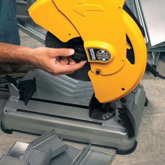 A close-up of a person adjusting a DEWALT D28715 yellow chop saw, used for cutting metal pieces. Several metal angles are visible around the saw base on a workshop floor.