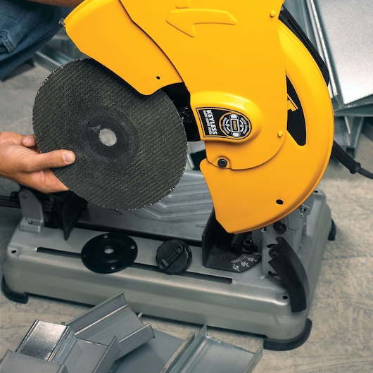 A person installing a cutting disc onto a yellow DEWALT chop saw, which is mounted on a gray base. Pieces of metal and metal profiles are visible near the tool.