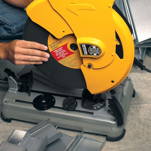 A person adjusting the blade of a DEWALT D28715 metal cutting saw. The saw features a yellow protective cover and is positioned on a grey base. Several pieces of metal are visible in the foreground.