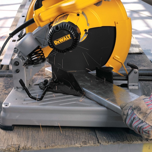 A DEWALT chop saw cutting through a metal piece, with sparks flying. The saw is mounted on a workbench and operated by a gloved hand.