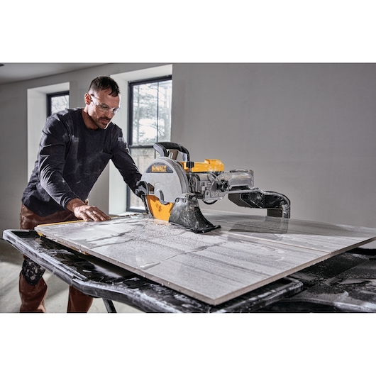 A person using a DEWALT D36000 tile saw to cut a large tile on a worktable in a room with two windows.