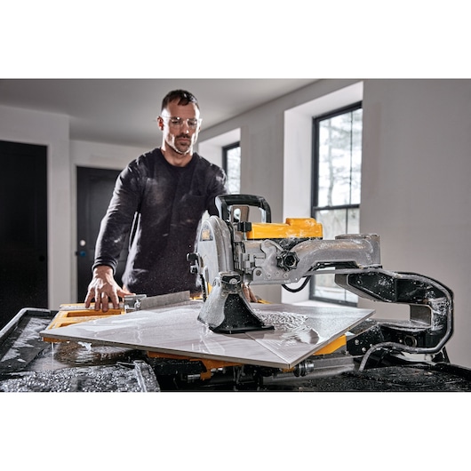 A person operating a DEWALT D36000-QS wet tile saw in a well-lit room, cutting a large tile with water splashing on the work surface.