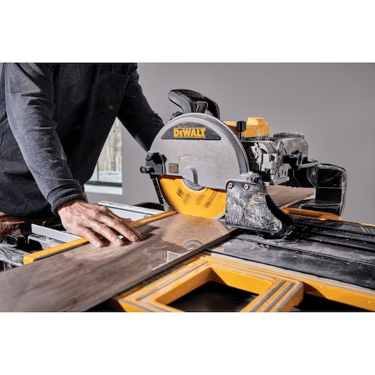 A person is cutting a large tile with a DEWALT tile saw. The saw blade is spinning and the person is guiding the tile carefully with their hand.