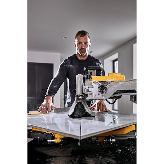 A person using a DEWALT tile cutting machine (SKU: D36000) to cut large ceramic tiles in an indoor setting. The image shows water and dust as part of the tile cutting process.