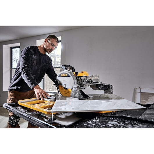A person operating a DEWALT D36000-QS wet tile saw to cut a large piece of tile indoors, with water running on the table for dust control. The person's face is blurred for privacy.