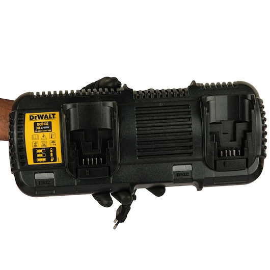 A DEWALT DCB132 dual battery charger for XR Li-Ion batteries, shown held by a gloved hand. The charger has two charging slots and a yellow label detailing specifications and warnings.