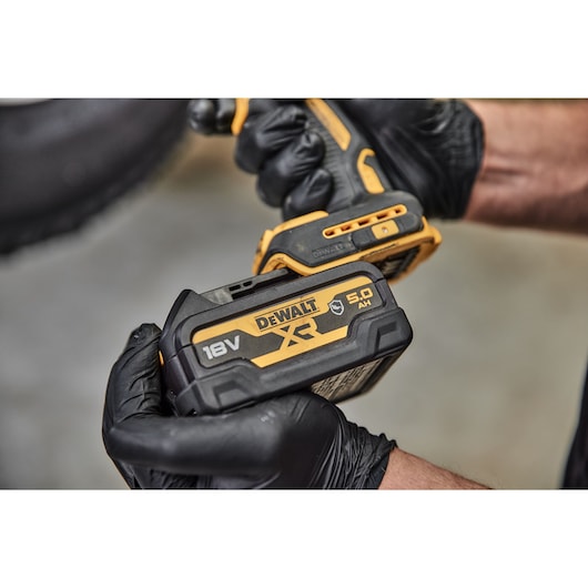 A close-up view of a person wearing black gloves holding a DEWALT cordless power tool with an attached DCB184G 18V XR lithium-ion battery.