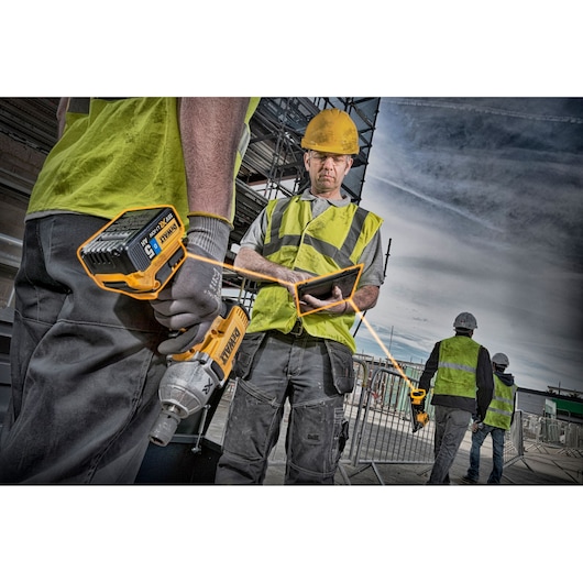 Construction workers wearing safety vests and helmets are seen at a work site. One worker is holding a DEWALT cordless power tool and another is using a tablet. Scaffolding and fencing are visible in the background.