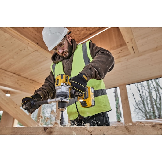 Person uses DEWALT drill with DCB1880 battery to drill wood. Wearing safety vest, gloves, and helmet at construction site.
