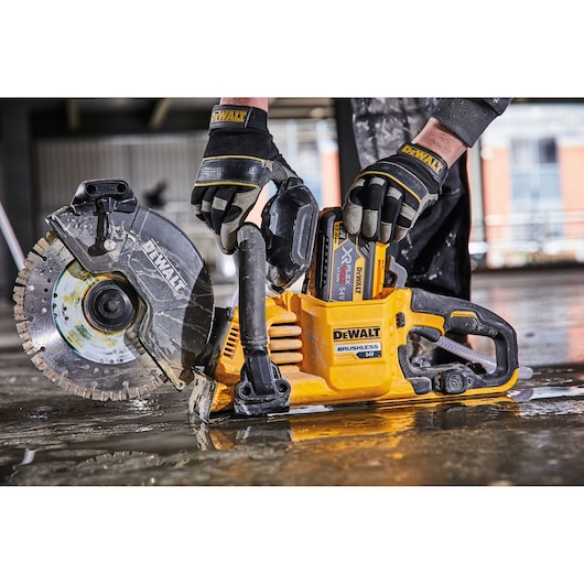 A person wearing protective gloves is handling a DEWALT 54V brushless cordless cut-off saw on a wet construction floor.