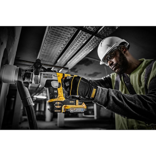 Worker drills into a wall with a DEWALT PowerStack 18V tool, wearing gloves and helmet on a construction site.