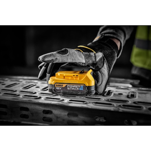 Gloved hand holds a DEWALT 18V POWERSTACK battery on a metal surface in a workshop setting.