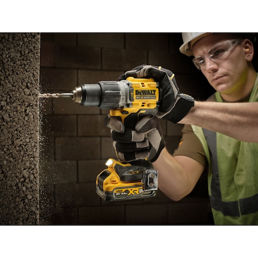 Gloved person uses a DEWALT cordless drill to bore into a concrete wall, with dust visible around the drill bit.