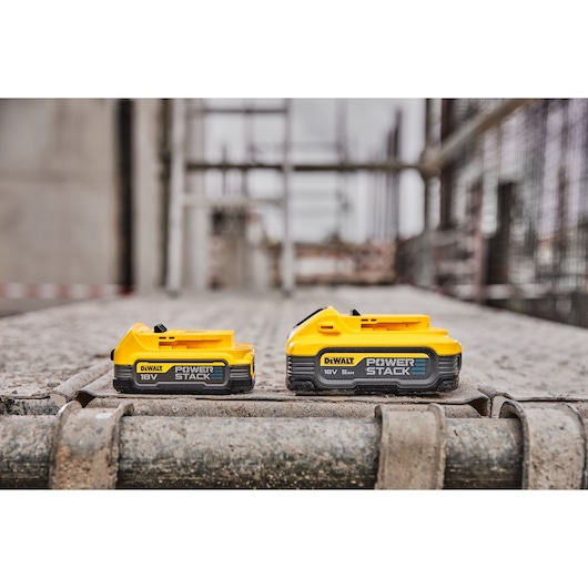 Two DEWALT 18V POWERSTACK batteries on a construction site surface, with a blurred industrial background.