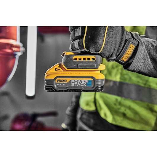 A gloved hand holds a DEWALT POWERSTACK 18V battery in a workshop setting, with safety gear visible.