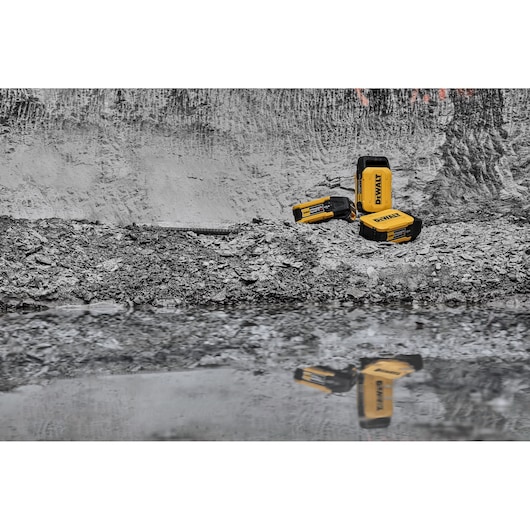 Three yellow and black DEWALT batteries are placed on rocky ground near a reflective puddle, with their reflection visible. The background is a textured gray wall, creating a contrast with the vibrant battery colors.