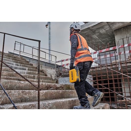 Worker climbs concrete stairs at construction site, holding a yellow DEWALT tool, wearing helmet and safety vest.