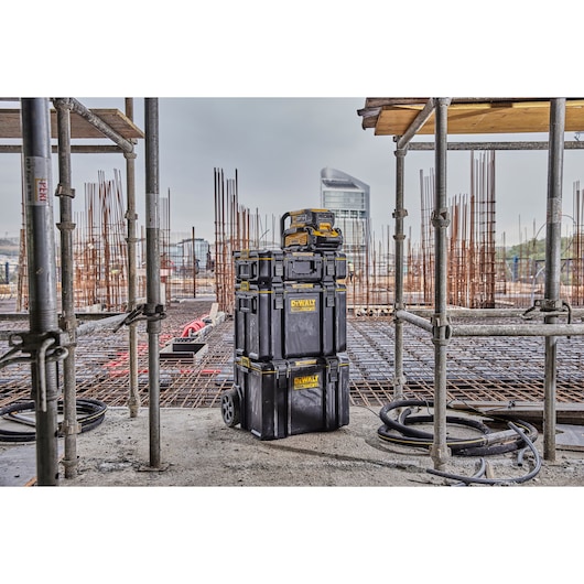 DEWALT stackable black tool storage boxes with yellow labels are shown on a construction site, surrounded by scaffolding and metal rods, with a modern building in the background.