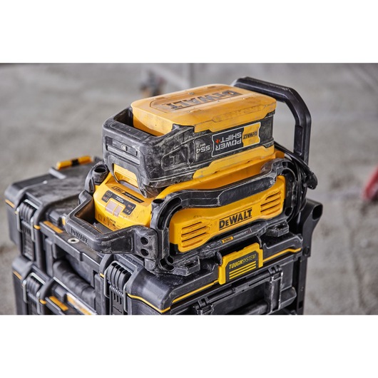 A close-up view of a DEWALT rechargeable battery and charging unit, both mounted on top of a stack of DEWALT ToughSystem storage boxes. The equipment appears rugged and suitable for worksite use.