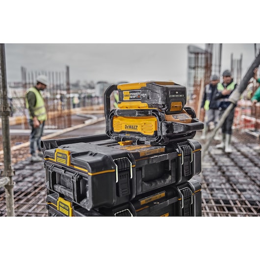 DEWALT PowerShift 554 battery and charging station placed on a stack of black and yellow DEWALT ToughSystem toolboxes at a construction site, with workers in the background.