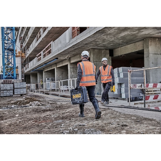 Two workers in orange vests carry DEWALT cases at a construction site with concrete building and safety barriers.