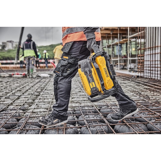 Worker carries a yellow DEWALT battery on a construction site with steel grid flooring.