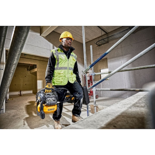 A construction worker wearing a yellow hard hat, high-visibility vest, gloves, and work boots is carrying a DEWALT portable air compressor (model DCC1054N) at a building site. The worker's face is blurred for privacy. The environment is under construction with scaffolding and unfinished walls.