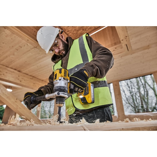 Person using a DEWALT cordless drill to bore wood, wearing safety gear and high-visibility vest on a construction site.