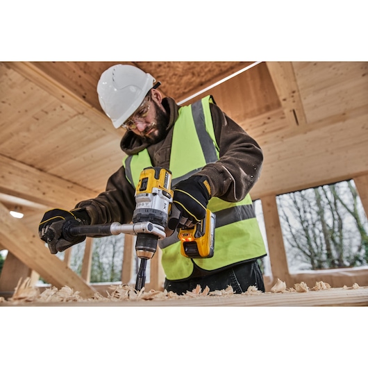 Person uses a DEWALT cordless drill on wood, wearing safety gear and a high-visibility vest at a construction site.