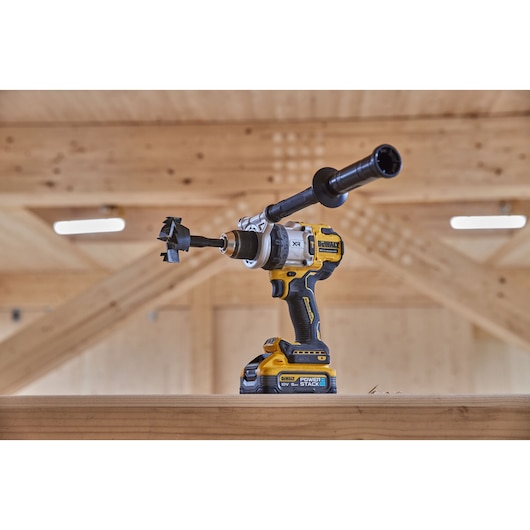 DEWALT cordless drill with side handle and spade bit, shown on a wooden beam in a workshop setting.
