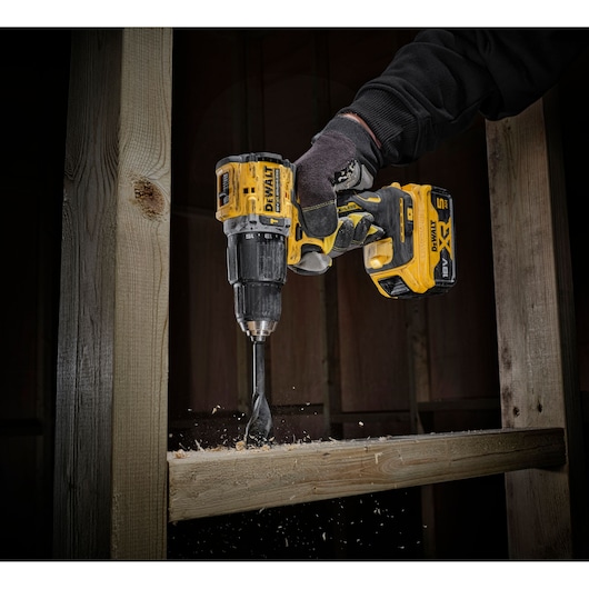 Gloved hand using a DEWALT cordless drill to bore a hole in a wooden beam, wood chips flying.