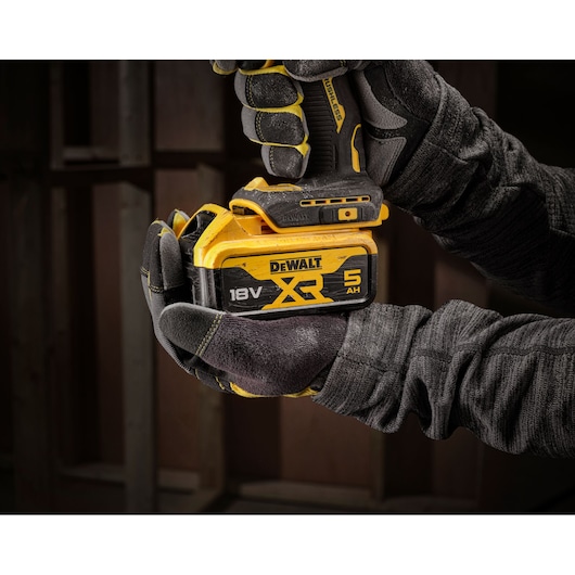 Gloved hands holding a DEWALT 18V XR 5Ah battery attached to a cordless tool in a workshop setting.